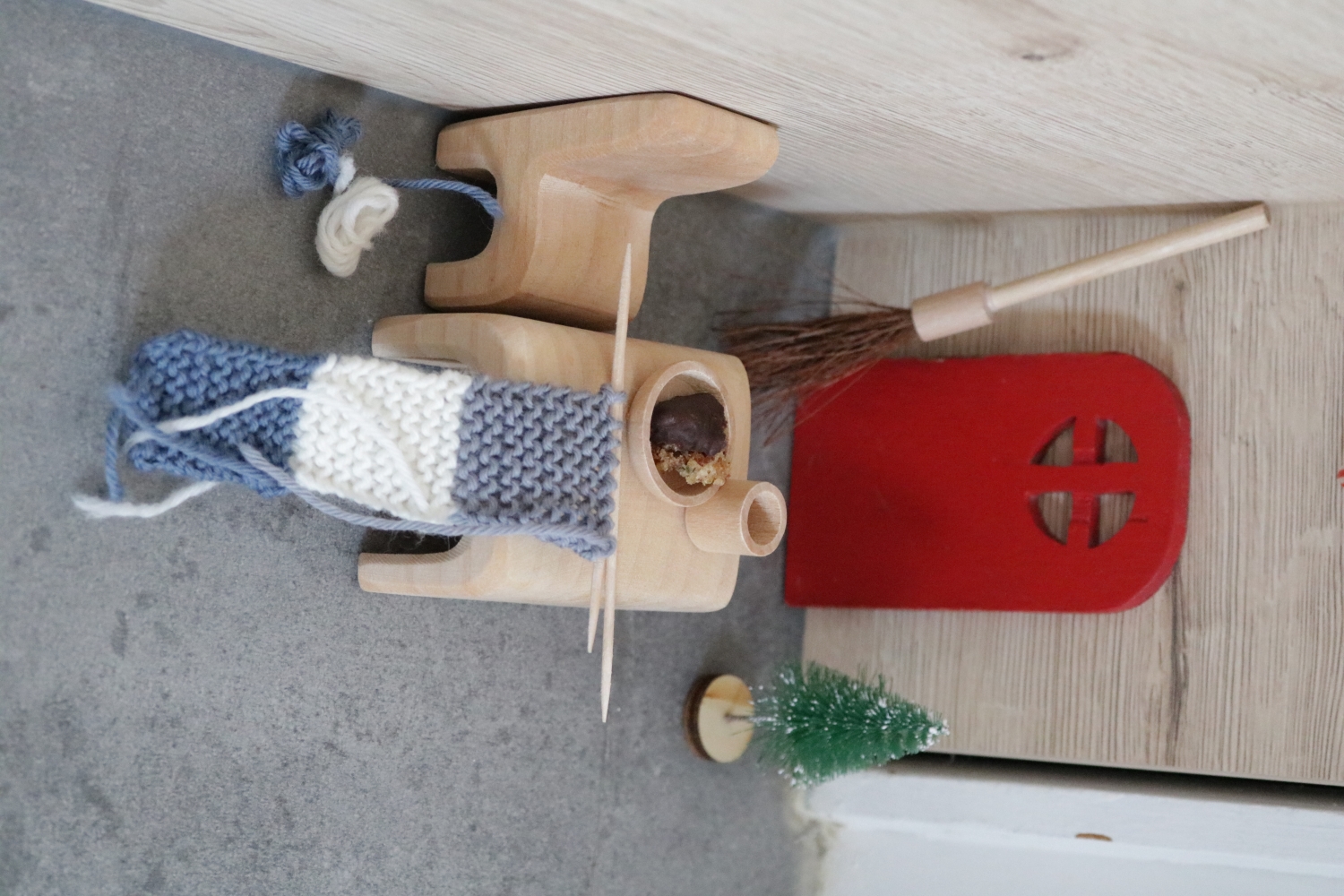 A doll-house table and chair in front of a little red door on the wall, on the table lies an unfinished scarf being knitted with toothpicks