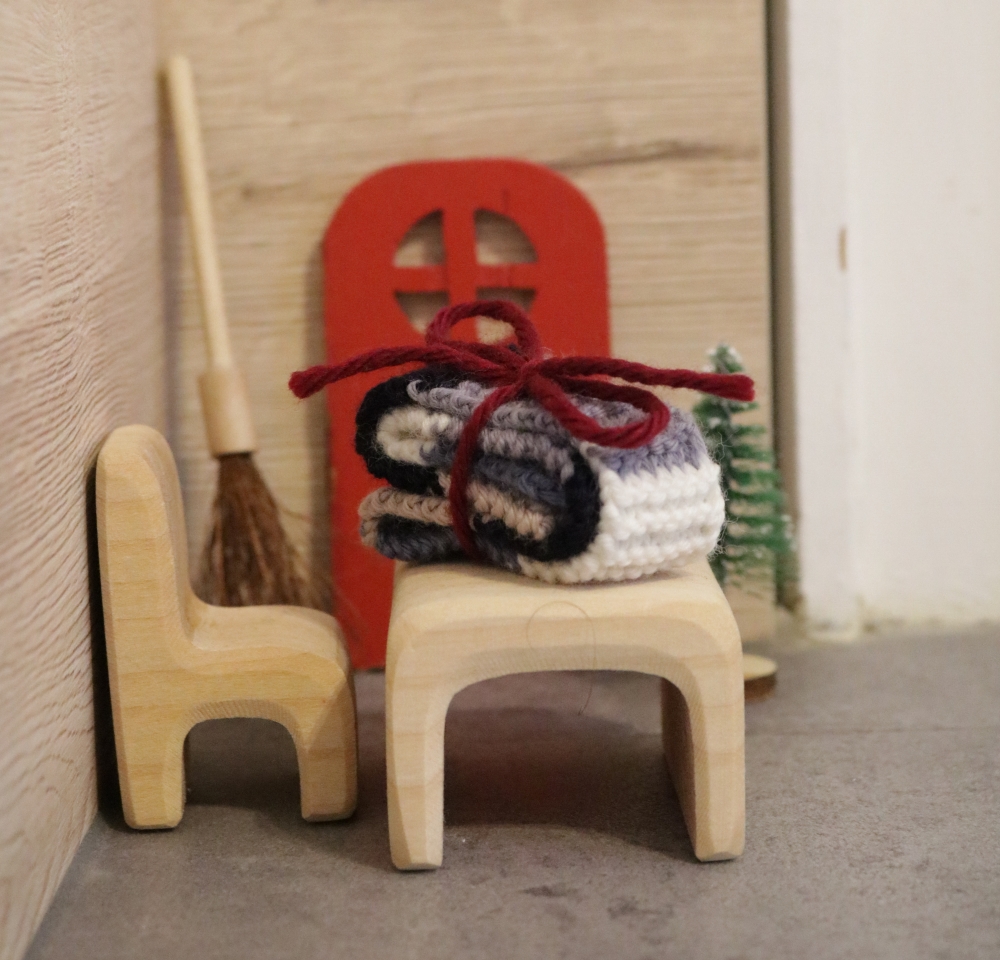 A doll-house table and chair in front of a little red door on the wall, on the table lies a tiny knitted scarf, folded and held together with a red bow