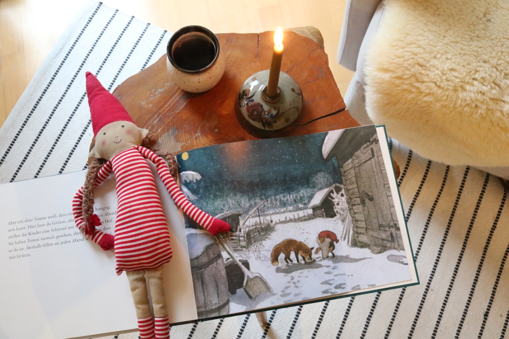 An open children's book with a winter illustration, an elf doll and a lit candle on a small wooden coffee table