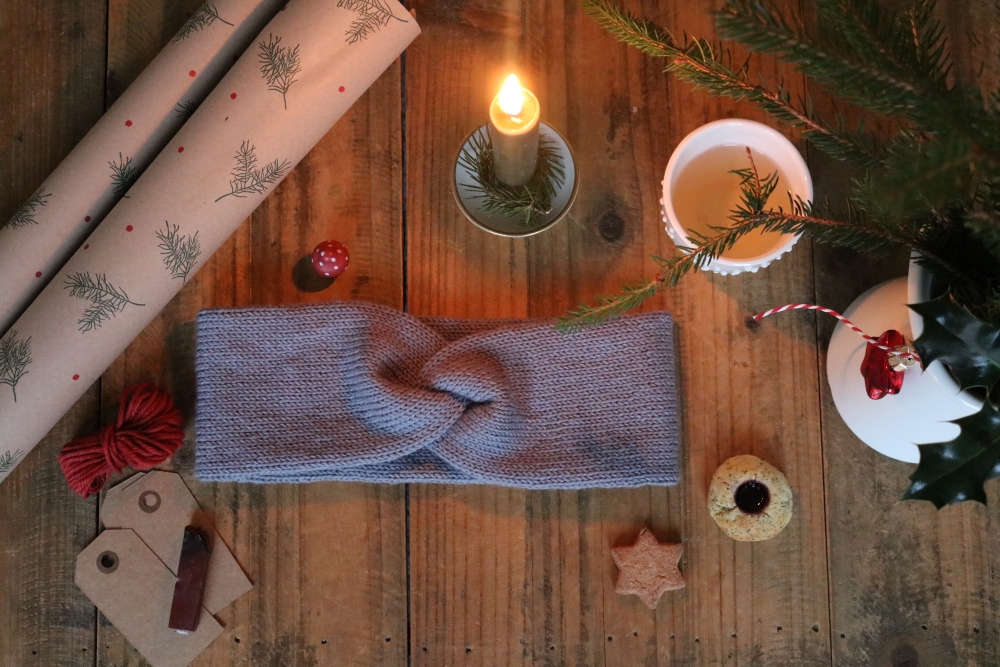 A grey knitted headband with a twist on a background of dark wood, around it some rolls of wrapping paper, a burning candle, fir branches and other Christmas decoration