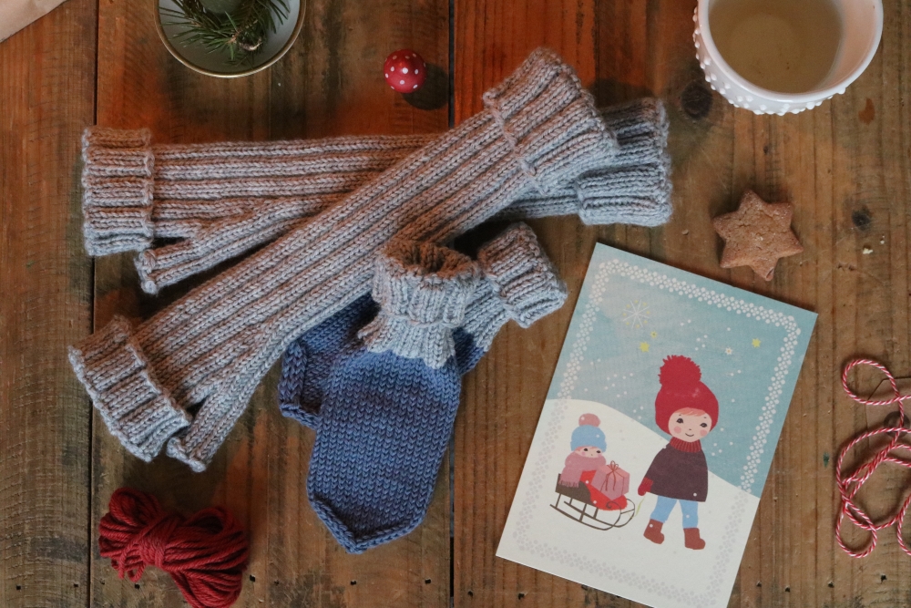 A pair of light-grey, long knitted wristwarmers and a pair of children's mittens in blue and grey on a  background of dark wood, next to them a card with a winter illustration and other Christmas decoration