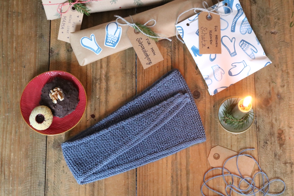 A small knitted scarf in blue on a background of dark wood, next to it some wrapped gifts, a burning candle and other Christmas decoration