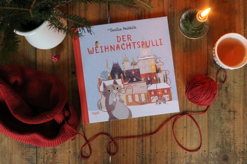 The German edition of the children's book A Christmas Sweater for Nina on a background of dark wood, next to it an unfinished knitted piece and a ball of yarn in dark red, a burning candle, a cup of tea and a few fir branches