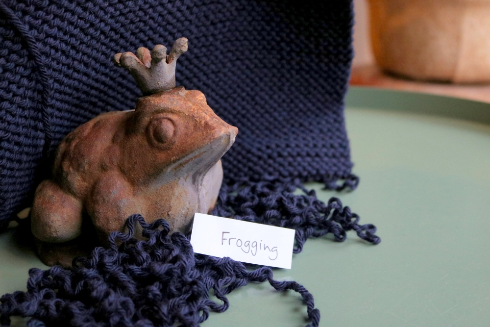 Ein Strickstück in Dunkelblau, davon kommend wieder aufgetrenntes Garn; inmitten des Garns ein Frosch aus Terracotta und ein kleines Stück Papier mit dem handgeschriebenen Wort Frogging darauf