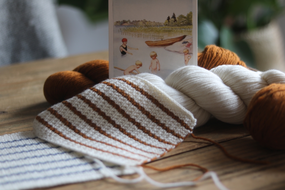 A knitted gauge swatch with stripes in white and terra on a wooden table. Next to it skeins of yarn in the same colours and a summery watercolour painting