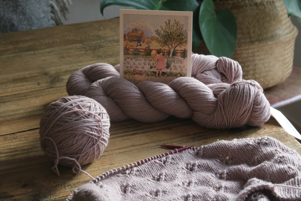 An unfinished knitted piece in beige on a wooden table. Next to it skeins of yarn in the same colour and a summery watercolour painting