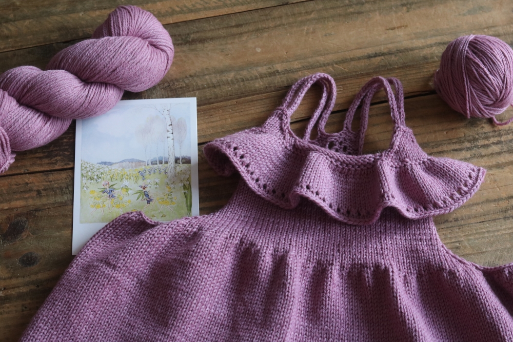 A knitted children's top with ruffles in powdery pink with a skein and a ball of yarn in the same colour, in the middle a small watercolour painting