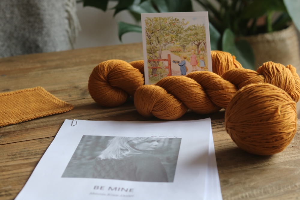 A printed knitting pattern on a wooden table. Next to it a ball of yarn ans some skeins in a caramel colour as well as a small summery watercolour painting